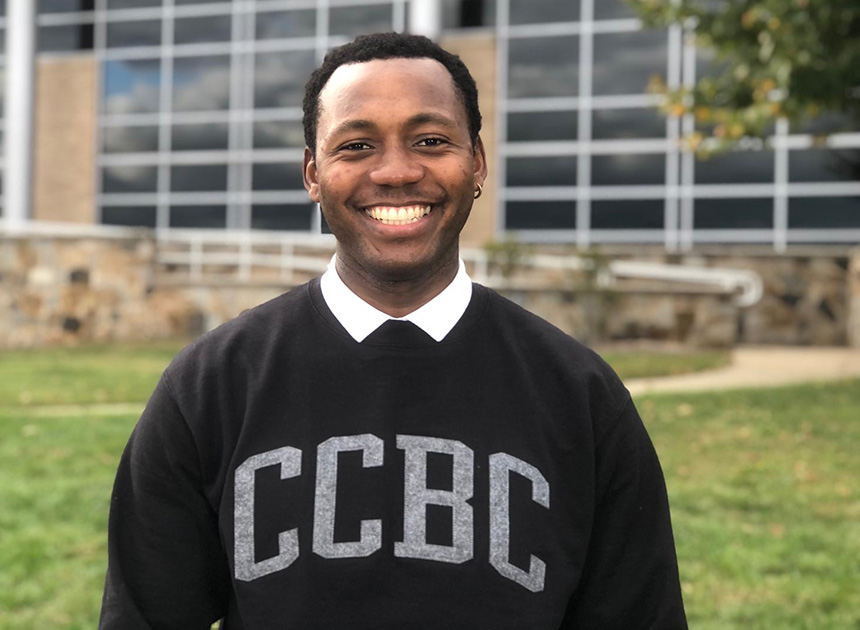 Student wearing a CCBC shirt smiles infront of a biulding on a sunny day