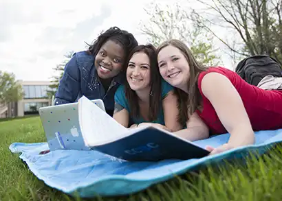 Three students are laying on a blanket studying outside