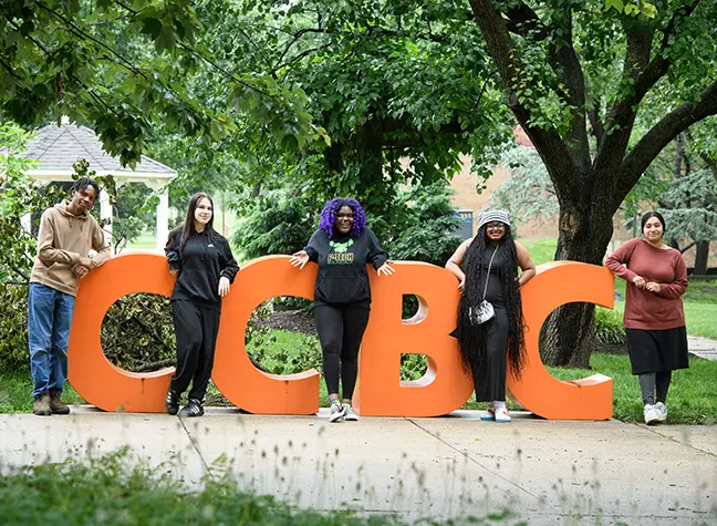 Five students stand between big orange letters that read "CCBC" on the CCBC Dundalk campus.
