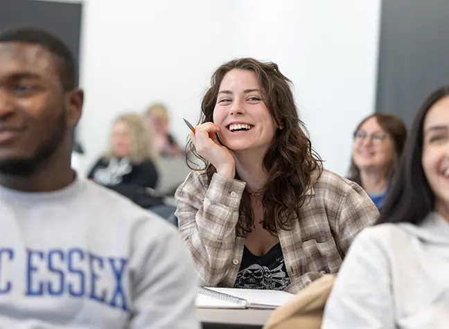 A student smiles cheerfully with a pencil in her hand surrounded by other students in a classroom.