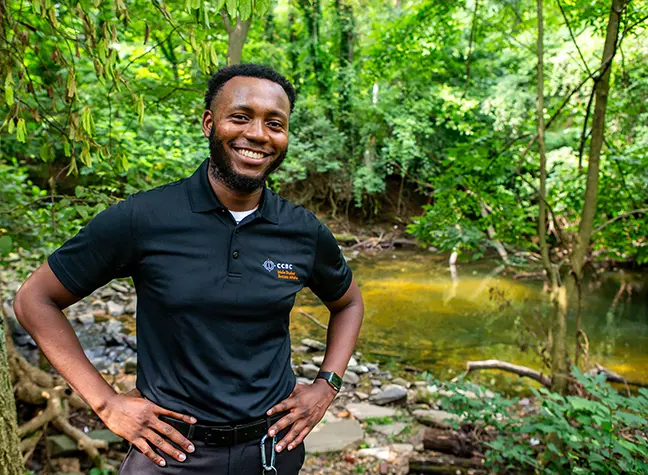 A teacher stands in front of a stream surrounded by green foliage.