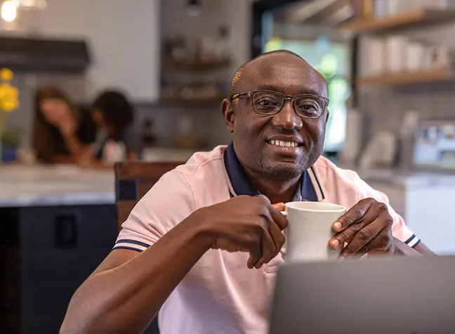     Man holds a cup of coffee as he sits in front of his laptop at home