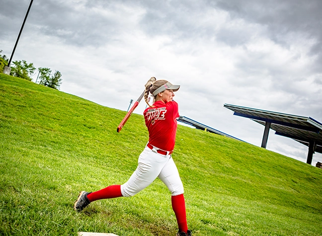 A softball player practices her throw on the softball field in front of a "Cardinals" banner.