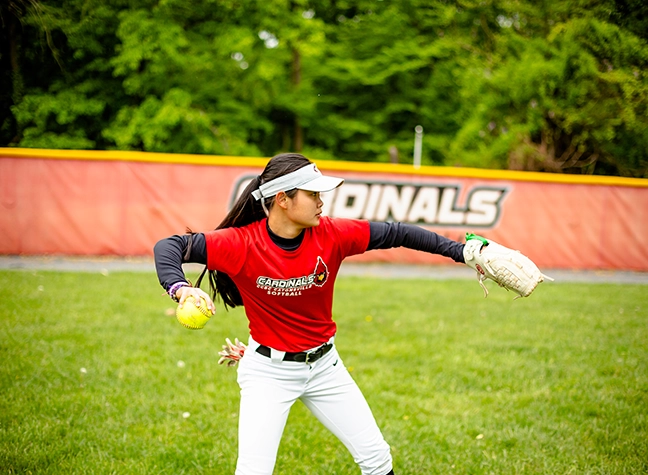A softball player practices her throw on the softball field in front of a "Cardinals" banner.