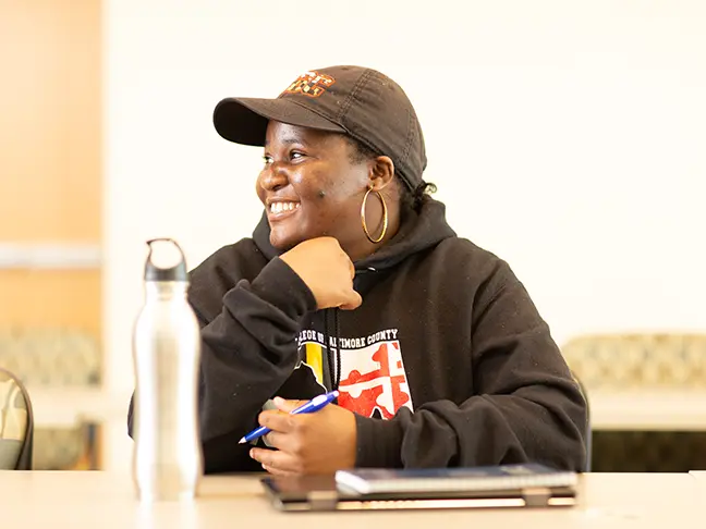 A student with a black baseball cap sits at her desk in a classroom and smiles to the left