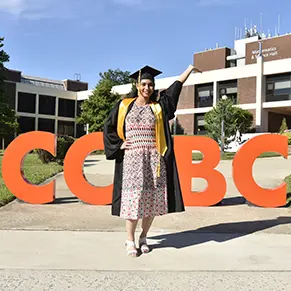 Chitpreet Kaur in her graduation gown standing in front of four oversized orange letters that spell out "CCBC"