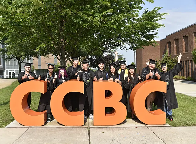 A group of students participating in CCBC's Commencement Ceremony gather around large orange letters that read, "CCBC."