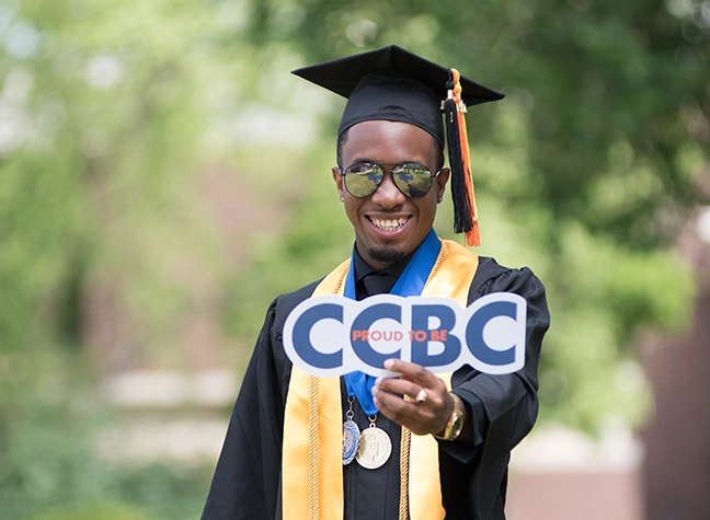 CCBC graduate wearing cap and gown, holding a proud to be CCBC sign
