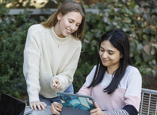 Two female students sitting outside looking over a tablet