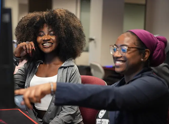 A woman points to a computer screen as another woman looks and laughs.