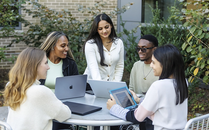 A group of THRIVE students sitting outside of an academic building, helping each other with projects on their laptops.