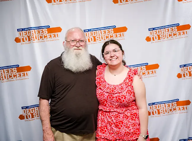 A father and daughter stand in front of a backdrop that reads "Degrees to Succeed."