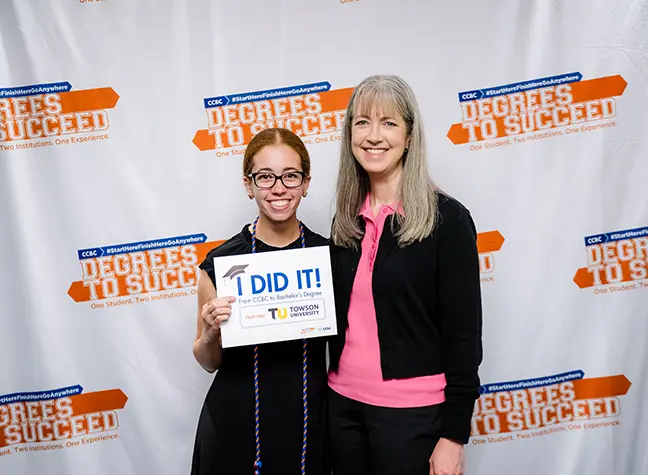 A degrees to succeed participant recently graduated from CCBC and is headed to Towson University. She crouches down with her child while holding a Degrees to Succeed certificate that reads "Towson."