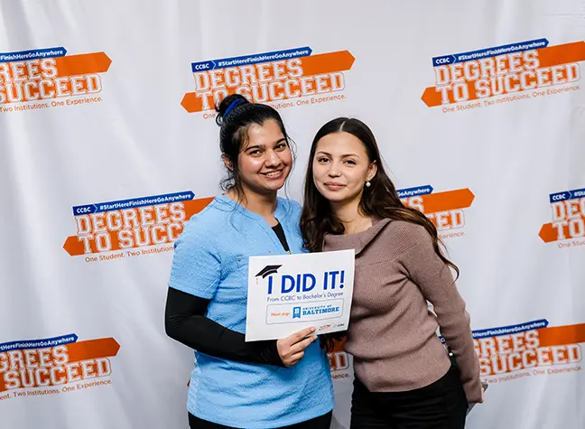 A CCBC student stands with her friend at the Degrees to Succeed Commencement ceremony holding a sign that reads "I did it!" with the University of Baltimore Logo on it.