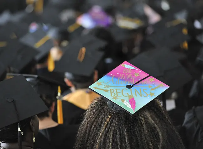 The back of a student's graduation cap reads, "And so the adventure begins!"