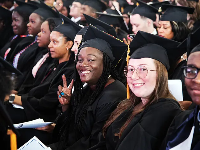 A graduate attending the CCBC Commencement ceremony is seated and holding up the peace sign.